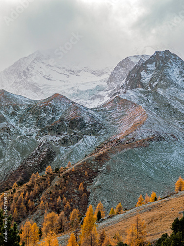 Amazing views over an high alpine valley and snow covered mountains. Orange and yellow coloured autumn and fall trees line the valley sides as the peaks rise above the clouds in this wilderness.