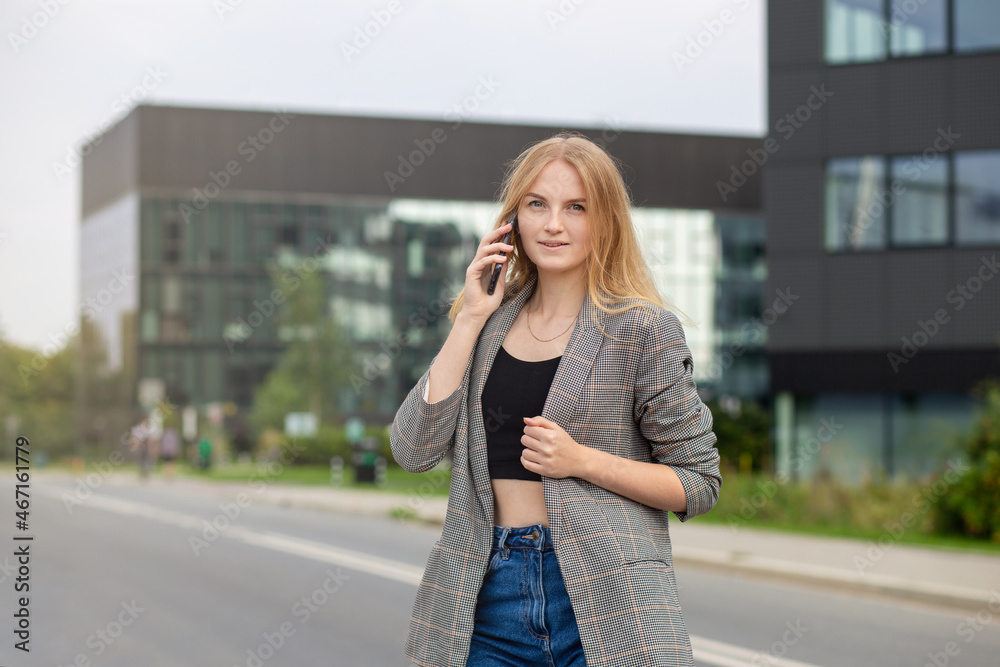 Portrait of young happy blond woman with phone walking on the street in the city. Technology or people concept. Excited girl smiling on bulding background.