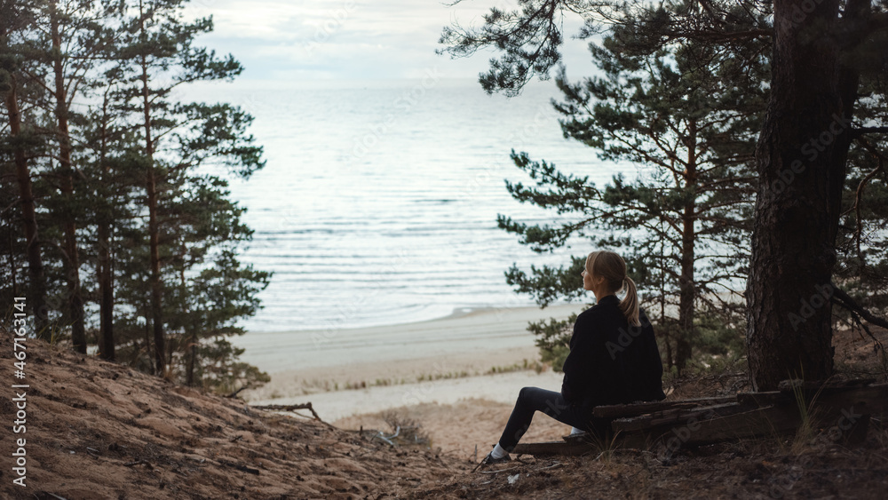 Portrait of a Young Beautiful Blond Woman in a Romantic Nature Atmosphere. Girl is Dressed in Black and is Sitting Alone in a Forest. She is Looking at a Sea Landscape.