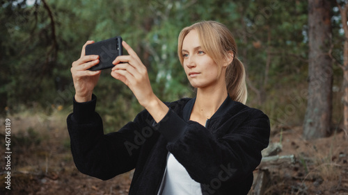 Portrait of a Young Beautiful Blond Woman in a Romantic Nature Atmosphere. Girl is Dressed in Black and is Taking a Picture on Her Mobile Phone. She Sits in a Pine Forest.