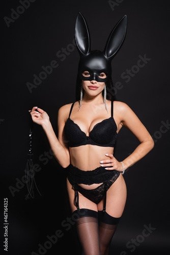seductive woman in underwear and bunny mask holding flogging whip while posing with hand on hip on black