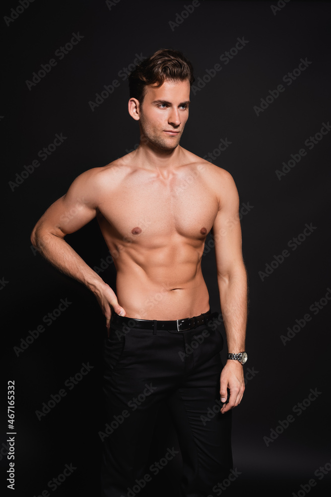 shirtless man in trousers looking away while standing with hand on hip on black