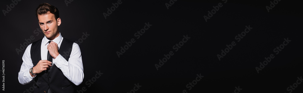 elegant and confident man adjusting tie while looking at camera isolated on black, banner