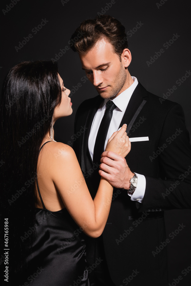 young man in elegant suit holding hand of stylish brunette woman isolated on black