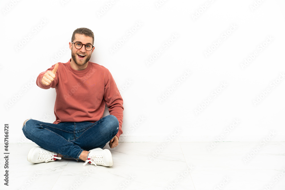 Young handsome man sitting on the floor with thumbs up because something good has happened Young handsome man sitting on the floor with thumbs up because something good has happened