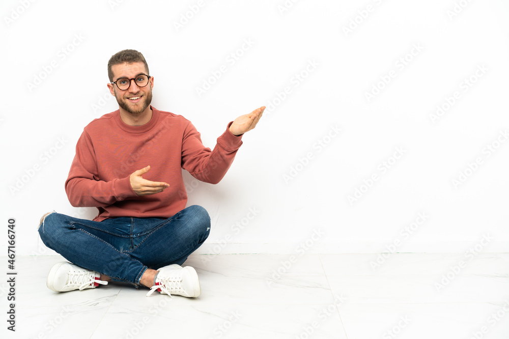 Young handsome man sitting on the floor extending hands to the side for inviting to come Young handsome man sitting on the floor extending hands to the side for inviting to come