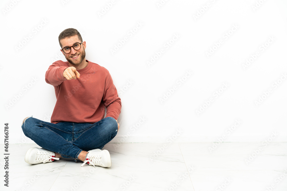 Young handsome man sitting on the floor points finger at you with a confident expression