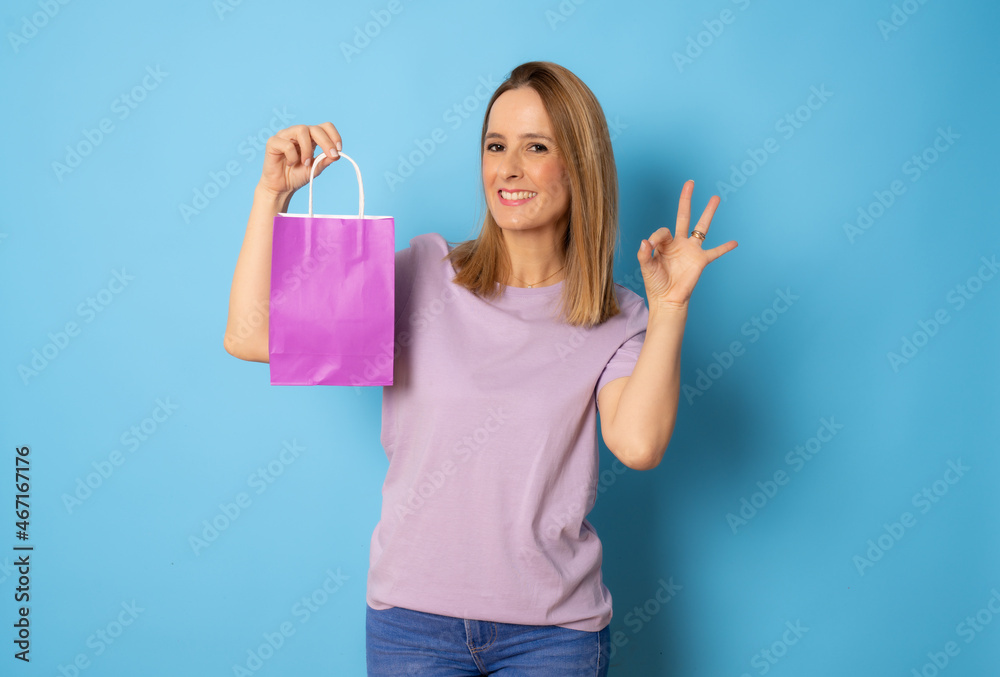 Young smiling woman in casual t-shirt holding a purple bag with okay sign isolated over blue background. Young smiling woman in casual t-shirt holding a purple bag with okay sign isolated over blue background.