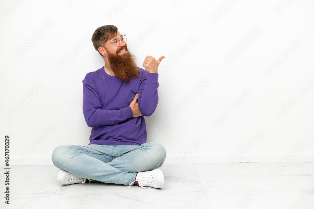 Young caucasian reddish man sitting on the floor isolated on white background pointing to the side to present a product Young caucasian reddish man sitting on the floor isolated on white background pointing to the side to present a product