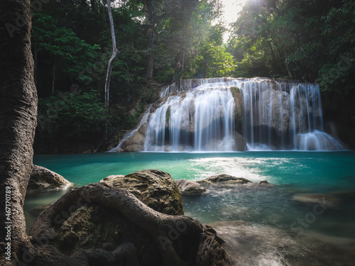 Scenic view of breathtaking Erawan Waterfall smooth flowing water and sunlight shine down to emerald green pond in lush rainforest with tree root foreground. Kanchanaburi  Thailand. Long exposure.