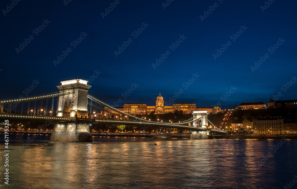 Night view of the old irradiated Chain Bridge in Budapest (Hungary), reflected in the river Danube. In the background you can see the historic houses of the city.