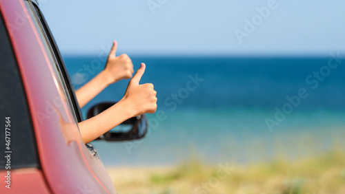 Two children's hands with a thumbs up gesture from the car window on the background of the sea and the beach. Empty white space, selective focus on the near hand.