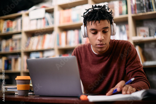 Young black student reads notes while e-learning on laptop in library.