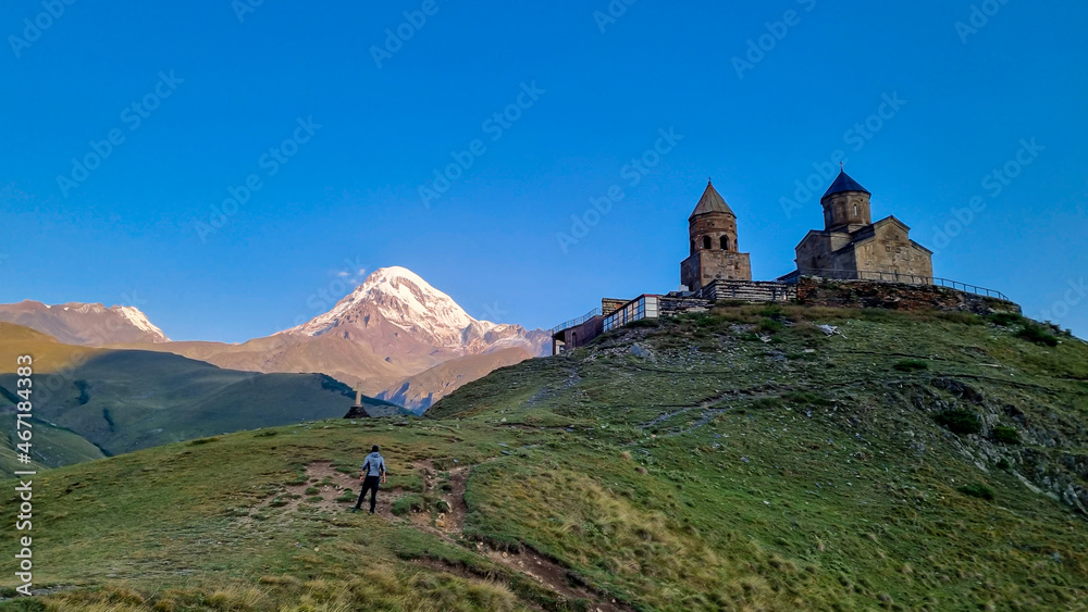 A man enjoying the view on Gergeti Trinity Church in Stepansminda, Georgia. The church is located on a high Caucasian mountain. Clear and blue sky above the church. In the back there are high mountain