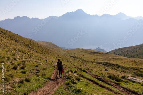 A woman walking on a narrow path towards in Caucasus, Georgia. High mountain chains in front. There slopes are overgrown with green grass. Cloudless sky. Exploring new places photo