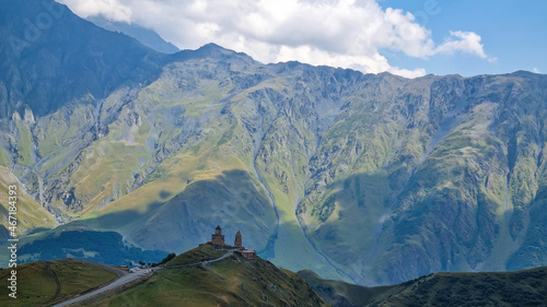 A distant view on Gergeti Trinity Church in Stepansminda, Georgia. The church is located on a high Caucasian mountain. Clear and blue sky above the church. In the back there are high mountain chains.