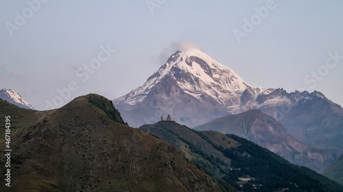 Distant view on Gergeti Trinity Church in Stepansminda, Georgia. The church is located on a high Caucasian mountain. Clear and blue sky above the church. Snow-capped Mount Kazbeg in the back. Daybreak