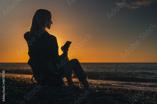 Young woman is sitting on a pebble beach by the sea with a mobile phone in her hands at sunset, a photo against the sun. Concept of happiness, travel, lifestyle