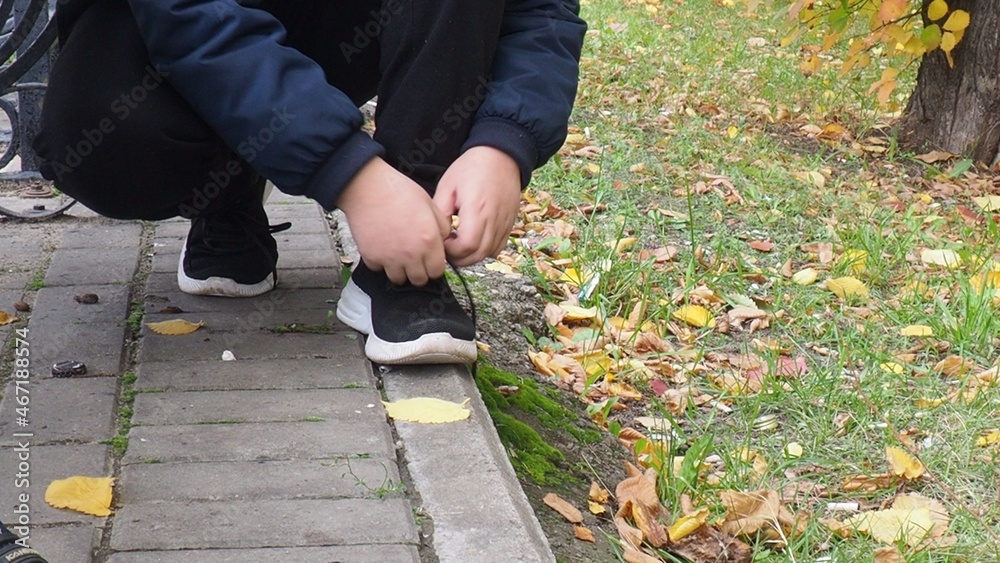 Closeup of unrecognizable sport boy tying sports shoes during evening run outdoors. a schoolboy teenager in a warm jacket walks in an autumn park with colorful foliage. Outdoor fun by any weather Closeup of unrecognizable sport boy tying sports shoes during evening run outdoors. a schoolboy teenager in a warm jacket walks in an autumn park with colorful foliage. Outdoor fun by any weather