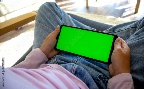 child at home sits in a chair using a smartphone with a green screen mockup in horizontal landscape mode. Using a mobile phone, browsing the Internet, watching content, videos, blogging, playing