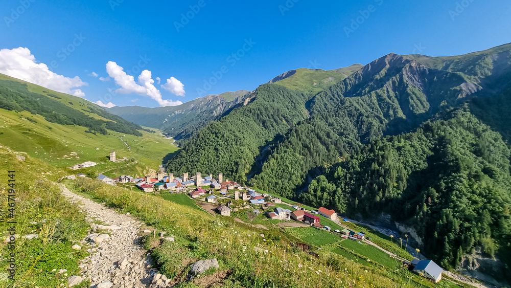 Panoramic view on Adishi, a mountain village, located in high Caucasus mountain chains. Few building on the bottom of the valley. Lush green pastures with a few cows grazing on them. Idyllic landscape
