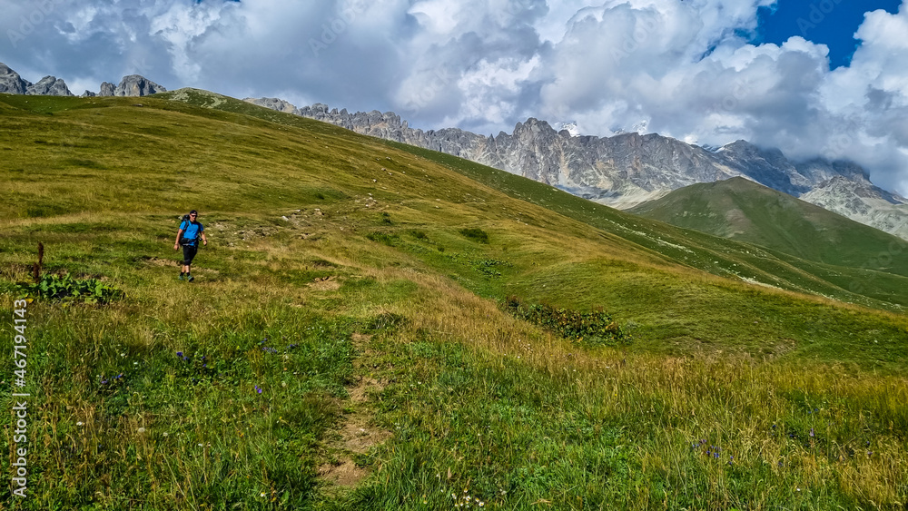 A man hiking along a narrow pathway in high Caucasus mountains in Georgia. There are high glaciers in the back. Thick clouds above the sharp peaks. Lush pastures on the sides. Barren peaks.
