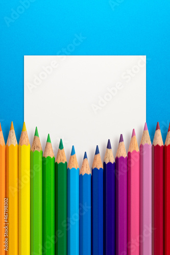 Creative layout made of color pencils on blue paper background with white paper card note. Flat lay. Education concept.