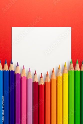 Creative layout made of color pencils on red paper background with white paper card note. Flat lay. Education concept.
