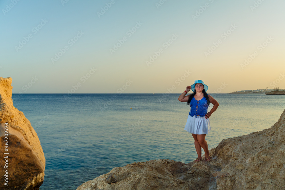 A young woman in a white skirt and a sun hat stands on the shore on the rocks against the backdrop of the sea at sunset.
