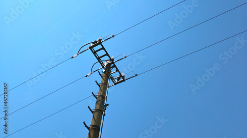 Detail of top part of low voltage pole of local electrical grid seen against blue sky. There are three lines of metal wire fixed to the pole with insulators and bridged by cable.