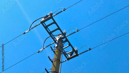 Detail of top part of low voltage pole of local electrical grid seen against blue sky. There are three lines of metal wire fixed to the pole with insulators and bridged by cable.