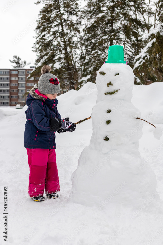 Small child making snowman, playing with snow on snowy cold winter day. Girl wearing snowsuit, having fun outdoors in park near house .
