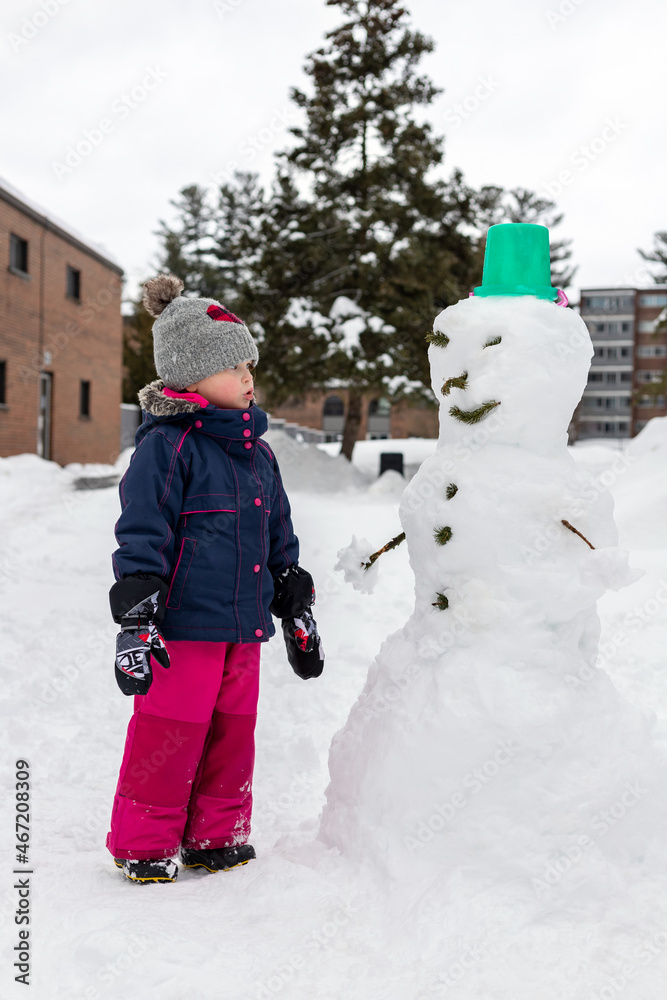 Small kid making snowman, playing with snow on snowy cold winter day. Girl wearing snowsuit, having fun outdoors.