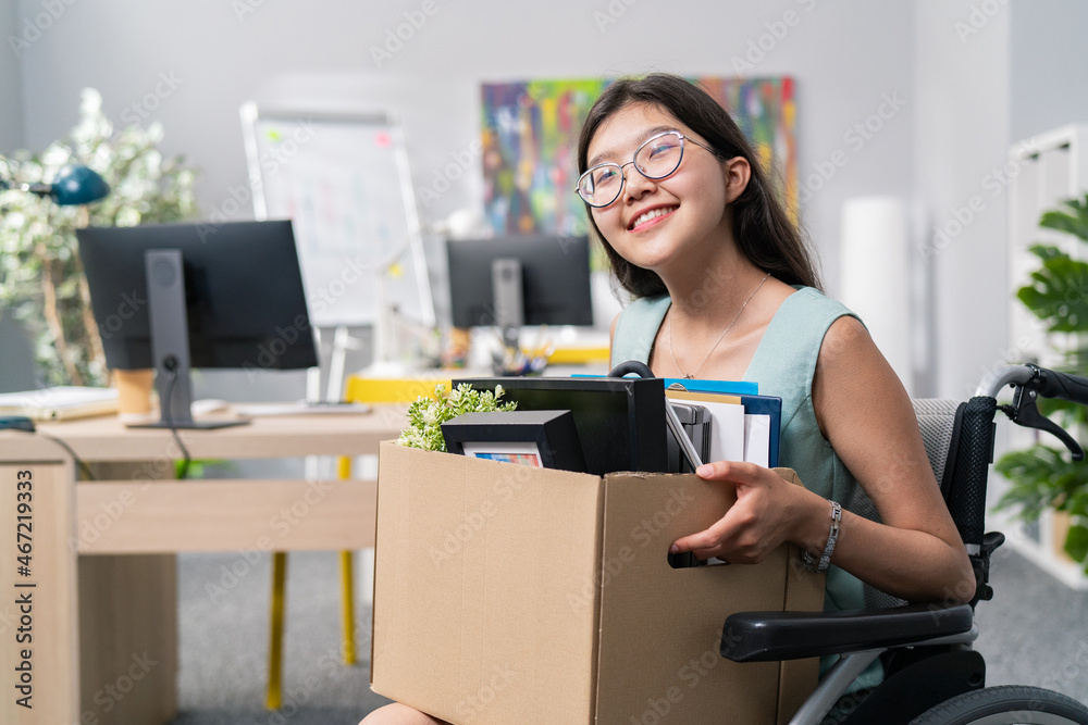 Positive smiling cheerful attractive young women with glasses is disabled rides in wheelchair and because of this gets promotion to another position, holds on lap cardboard box with things accessories