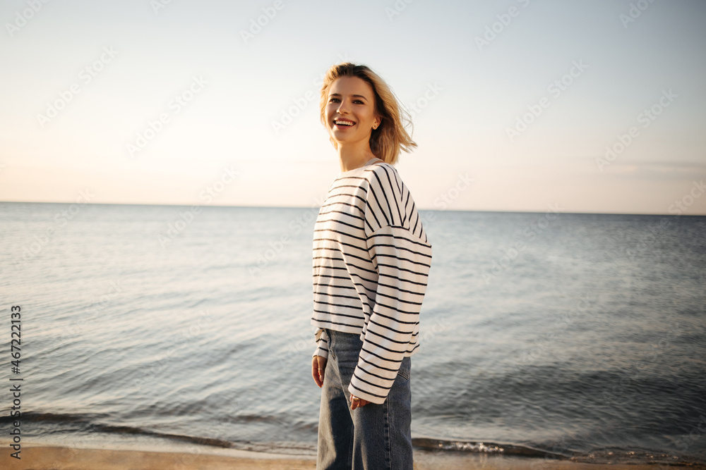 Happy pretty caucasian young woman stands on seashore smiling cute with teeth. Sophisticated blonde dressed in striped black and white blouse and dark jeans. Rest time concept. Happy pretty caucasian young woman stands on seashore smiling cute with teeth. Sophisticated blonde dressed in striped black and white blouse and dark jeans. Rest time concept.