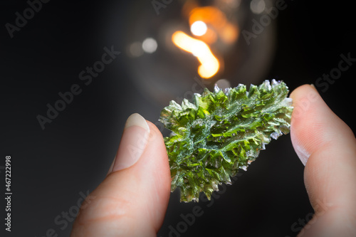 Rare green moldavite gem in human fingers with incandescent lamp on black background. Close-up of beautiful precious stone from meteoric glass with wrinkled mossy look. Raw mineral from South Bohemia. photo