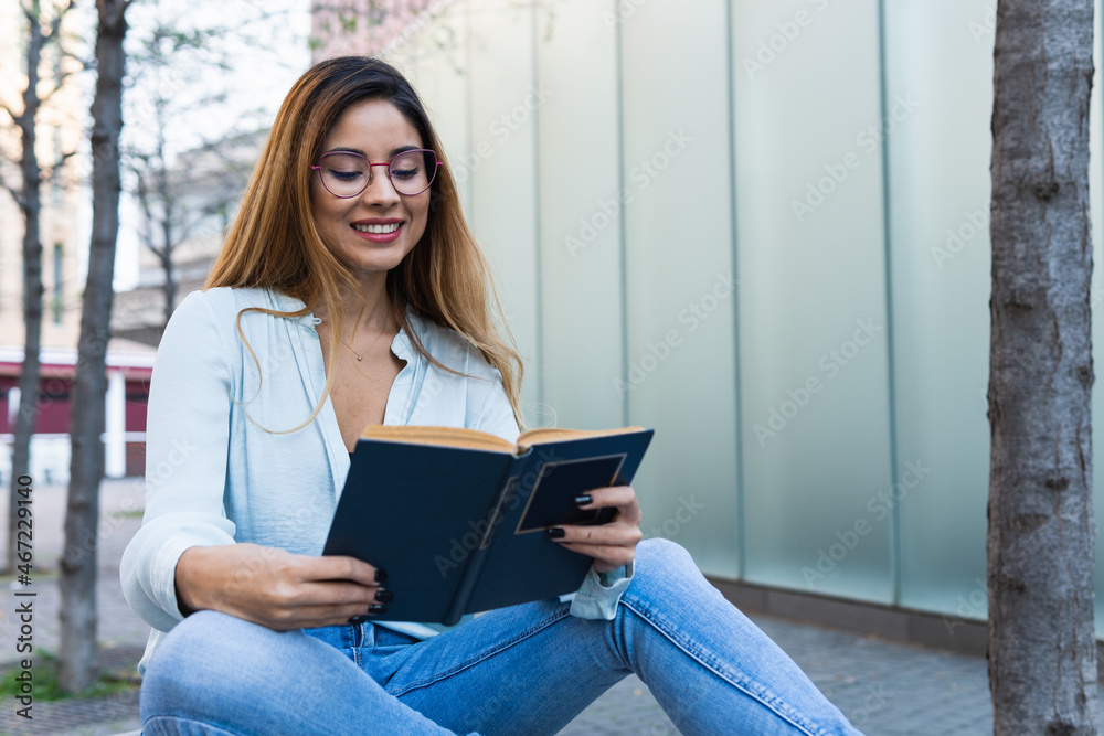 Fototapeta premium Positive optimistic brunette woman with glasses smiling while reading book outdoors. Student concept