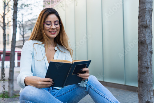 Positive optimistic brunette woman with glasses smiling while reading book outdoors. Student concept