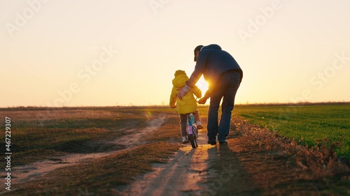 Father teaches a little kid girl to ride a child's bike on the road, in the fall, in spring. Happy family, childhood. Happy family, dad teaches his daughter, child to ride a bike in the park at sunset
