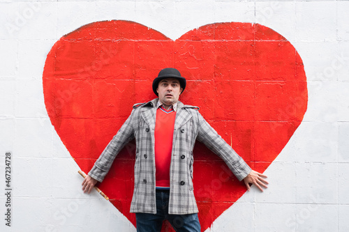 Emotional man in a hat and coat standing against a white wall against a background of a red heart. Love and relationship concept
