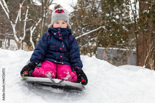 Little girl tobogganing downhill, having fun. Small happy child sledging in snow outdoors in winter. Little girl in hat and jacket sitting on sledge in cold weather.