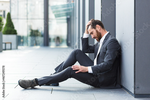 Sad depressed entrepreneur in formal suit worker man sitting near outdoors street near modern office business center. Upset male businessman lost job due financial crisis employee has problem. outside