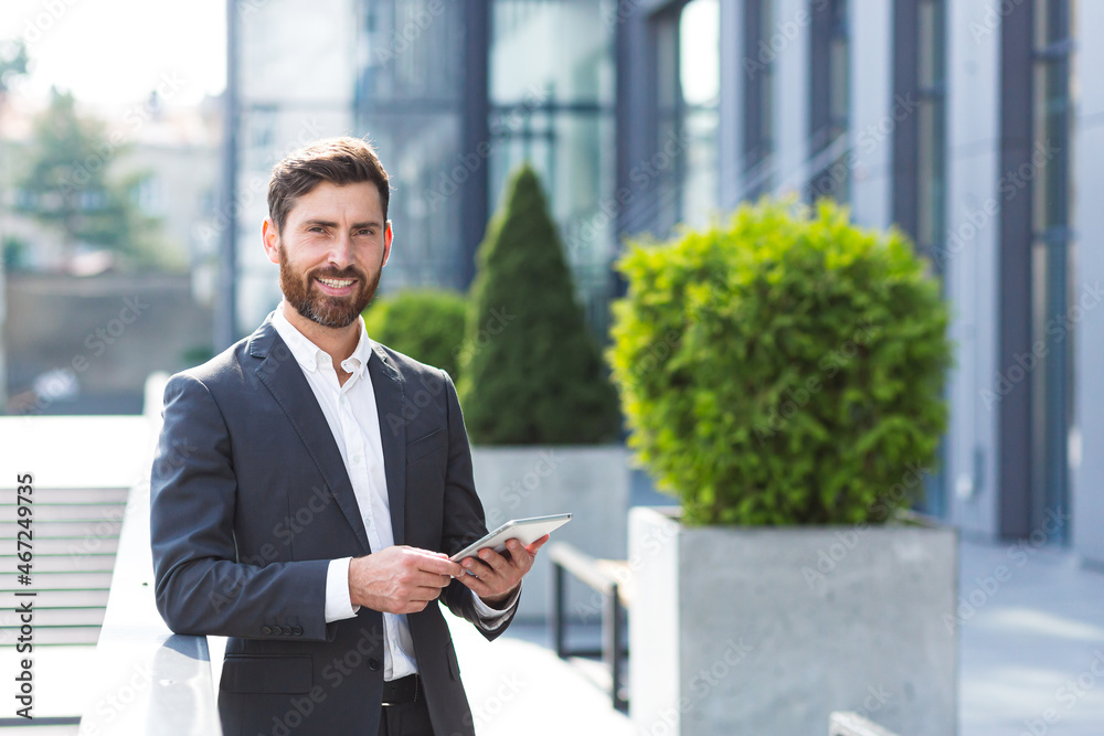 Cheerful and successful businessman works outdoors near the office holds a tablet, smiles reads good news
