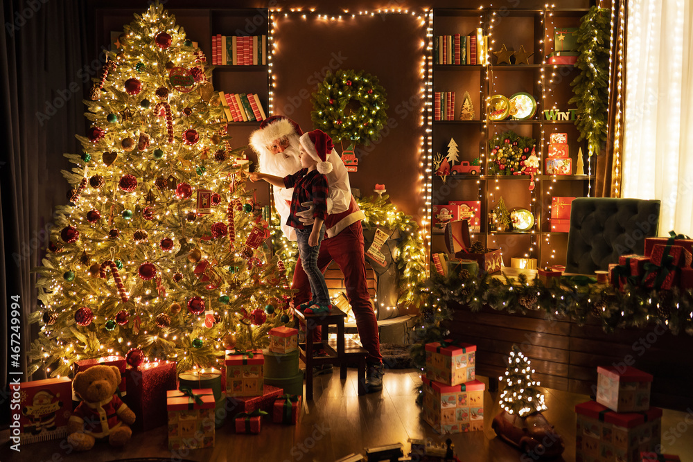 Full length of Santa Claus and his little assistant hangs balls decorate Christmas tree. 