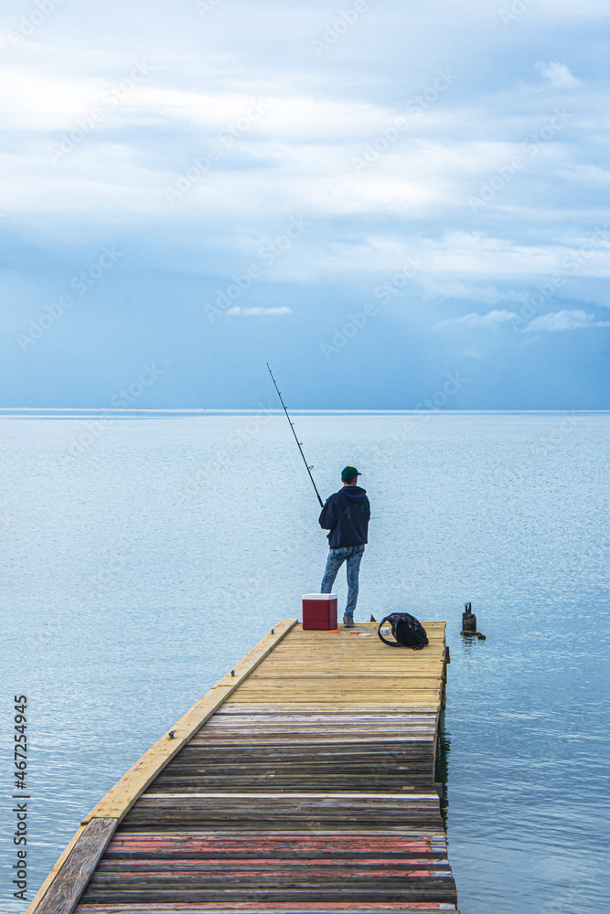 Young man fishing on dock with Caribbean Sea stretching to grey sky horizon.  Vertical format.  Salinas, Puerto Rico, USA.