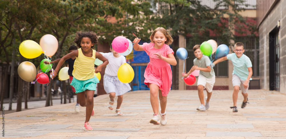 Obraz premium Multiracial group of cheerful tweenagers holding colored balloons in hands, running together on summer city street. Happy childhood concept