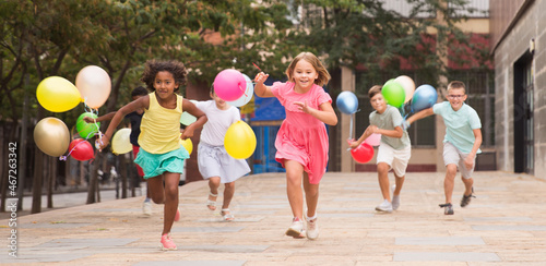 Multiracial group of cheerful tweenagers holding colored balloons in hands, running together on summer city street. Happy childhood concept