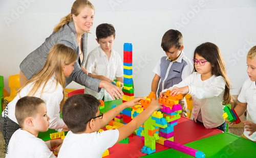 Young female teacher and happy schoolkids building castle from plastic toy blocks during lesson in classroom