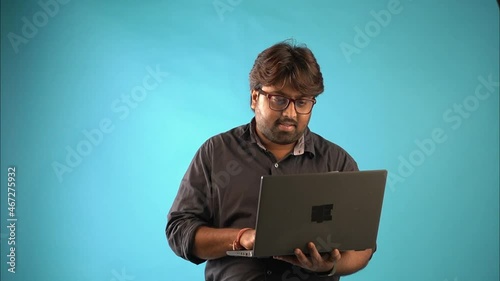 A Indian man in black shirt wearing glasses standing with laptop in an isolated blue background studio. photo