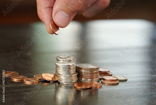 pile and stacking Thai coins on black table (selective focus) with reflection of sunlight, blurred background and senior man hand pick a coin up, copy space. concept for collecting and saving.